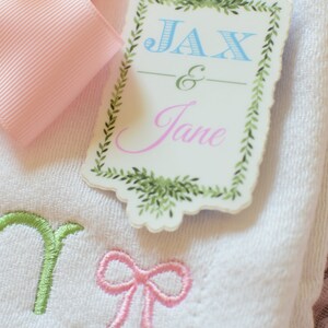 May include: A white towel with green, pink and blue embroidery. The towel has a tag attached to it that says "Jax & Jane" in a decorative font with a green leafy border.