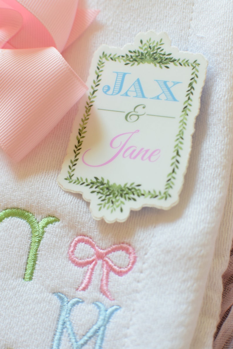 May include: A white towel with green, pink and blue embroidery. The towel has a tag attached to it that says "Jax & Jane" in a decorative font with a green leafy border.