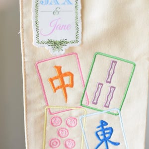 May include: Cream-colored fabric with embroidered Mahjong tiles in various colors. The tiles feature Chinese characters and symbols. A sticker at the top reads "JAX & Jane" in blue and pink, framed by green leaves.