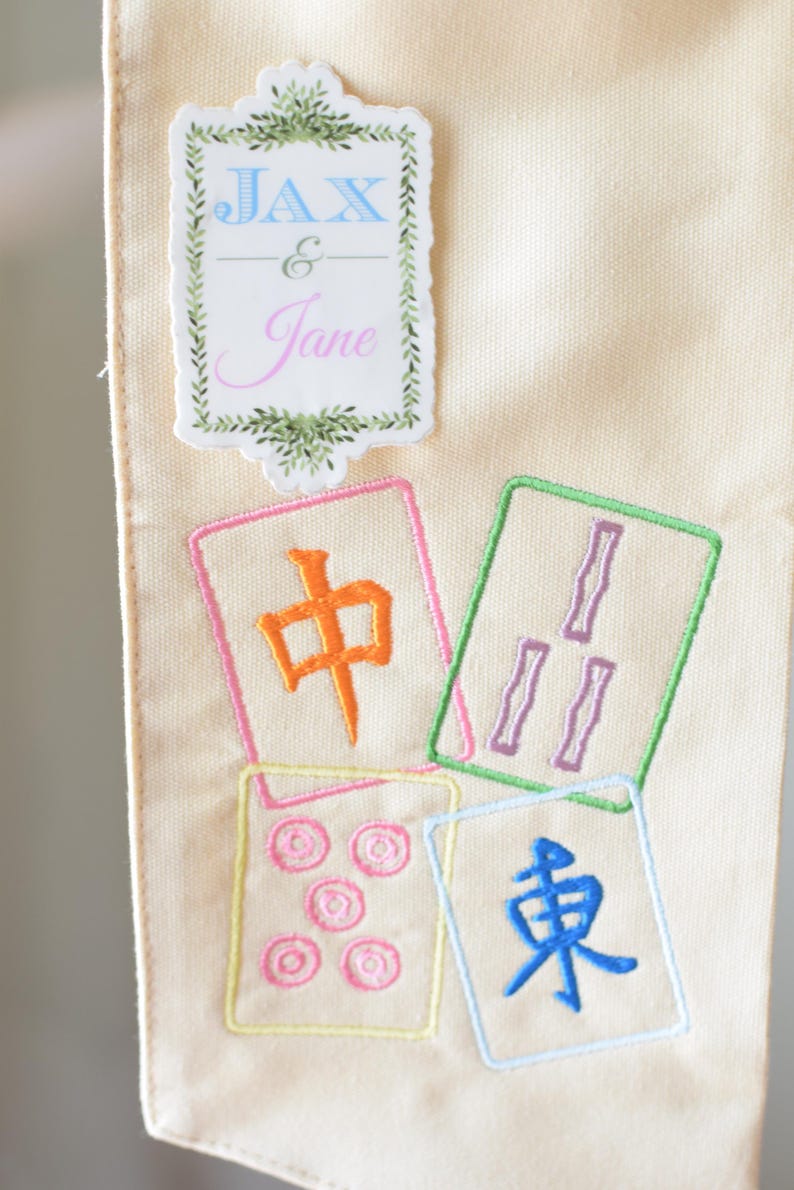 May include: Cream-colored fabric with embroidered Mahjong tiles in various colors. The tiles feature Chinese characters and symbols. A sticker at the top reads "JAX & Jane" in blue and pink, framed by green leaves.
