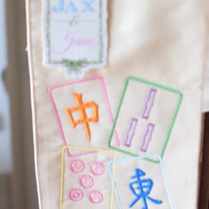May include: Cream-colored fabric with embroidered Mahjong tiles and a decorative label that reads "Jax & Jane". The tiles feature colorful Chinese characters and symbols, with a variety of colors including orange, purple, and blue.