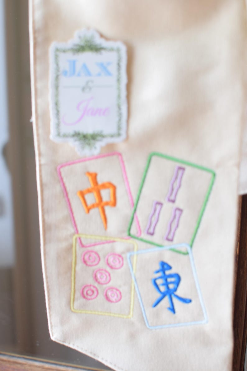 May include: Cream-colored fabric with embroidered Mahjong tiles and a decorative label that reads "Jax & Jane". The tiles feature colorful Chinese characters and symbols, with a variety of colors including orange, purple, and blue.
