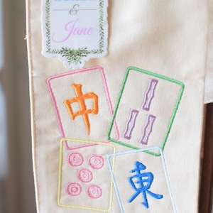 May include: Cream-colored fabric with embroidered Mahjong tiles in various colors. The tiles feature Chinese characters and symbols. A decorative label at the top reads "Jax & Jane" in blue and pink script.
