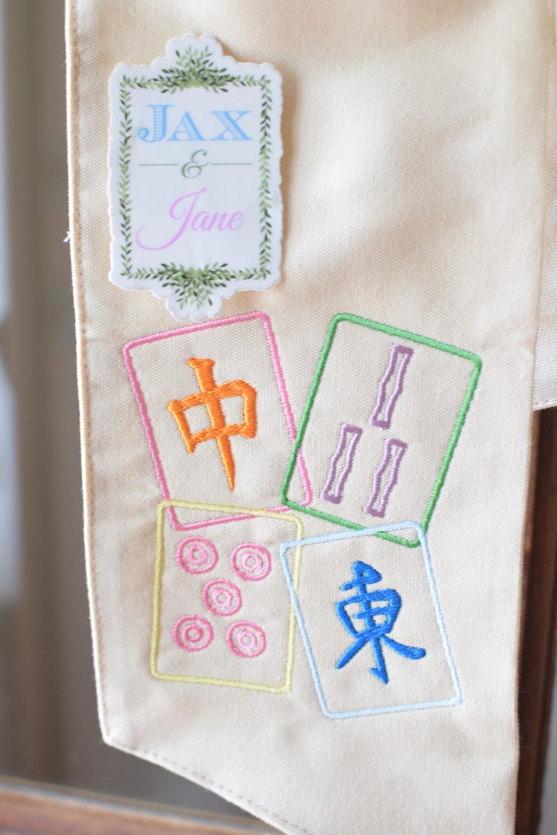 May include: Cream-colored fabric with embroidered Mahjong tiles in various colors. The tiles feature Chinese characters and symbols. A decorative label at the top reads "Jax & Jane" in blue and pink script.