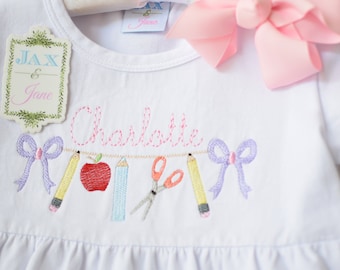 Könnte beinhalten: Weißes Kurzarmhemd mit rosa Stickerei des Namens "Charlotte" und einer Schnur mit Schulutensilien, darunter ein roter Apfel, Bleistifte, eine Schere und eine lila Schleife. Das Hemd hat einen Rüschen-Saum und ein Etikett mit der Aufschrift "Jax & Jane".