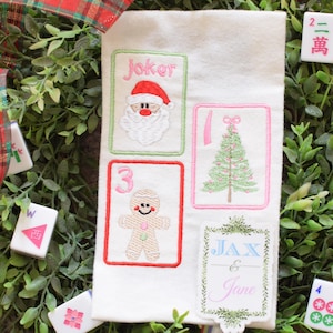 Könnte beinhalten: Ein weißes, weihnachtliches, besticktes Stoffstück mit vier Kartendesigns: ein Weihnachtsmann, ein Weihnachtsbaum, ein Lebkuchenmann und eine Karte mit dem Text "Jax & Jane". Der Stoff ist von Grün, rot-grün kariertem Band und Mahjong-Steinen umgeben.