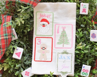 May include: A white Christmas-themed embroidered fabric with four card designs: a Santa, a Christmas tree, a gingerbread man, and a card with the text "Jax & Jane". The fabric is surrounded by greenery, red and green plaid ribbon, and Mahjong tiles.