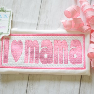 May include: White fabric with pink embroidered lettering that reads "I love mama" with a pink heart in place of the "o".