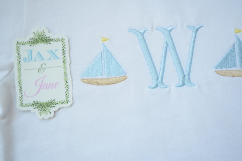 May include: A white embroidered baby blanket with two blue sailboats and the letter "W" in blue thread. A tag with the text "Jax & Jane" is in the upper left corner.