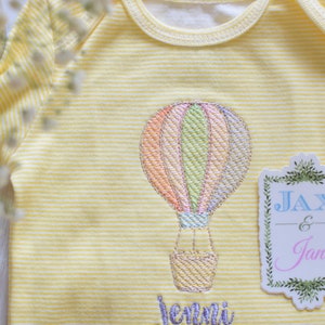 May include: Yellow striped baby onesie with an embroidered hot air balloon and the text "Jax & Jane" on a green and white tag.