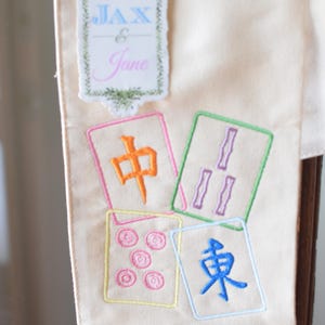 May include: Cream-colored fabric banner with embroidered Mahjong tiles and the names "Jax & Jane". The tiles feature colorful symbols and characters. The banner is likely used for decoration or a special event.