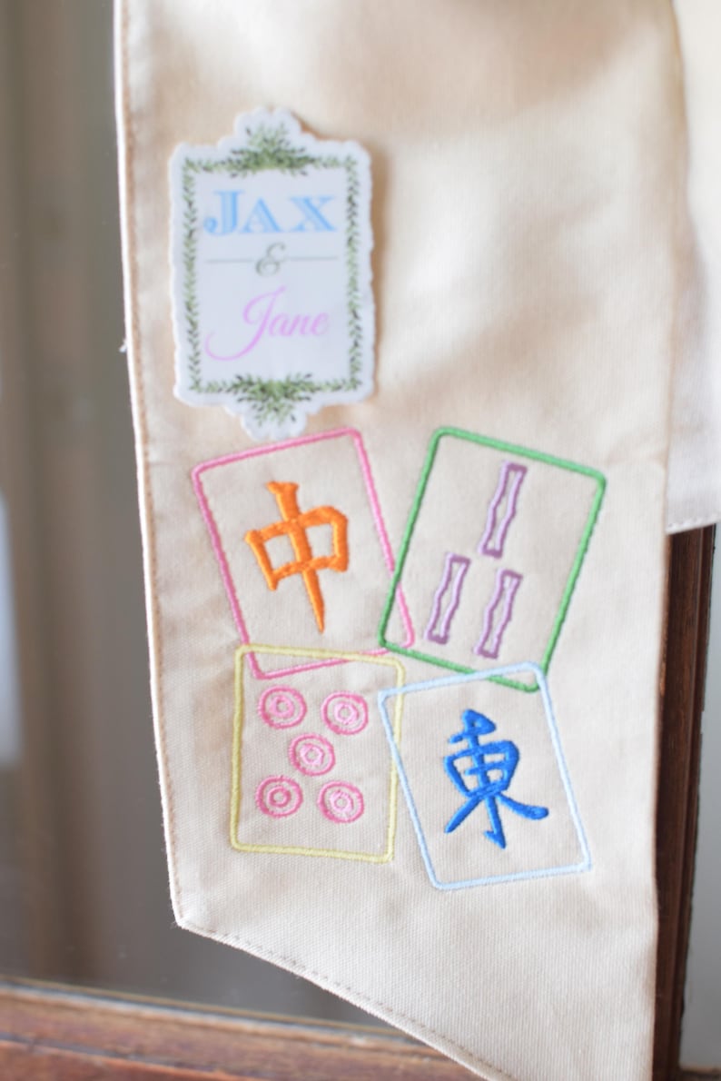 May include: Cream-colored fabric banner with embroidered Mahjong tiles and the names "Jax & Jane". The tiles feature colorful symbols and characters. The banner is likely used for decoration or a special event.