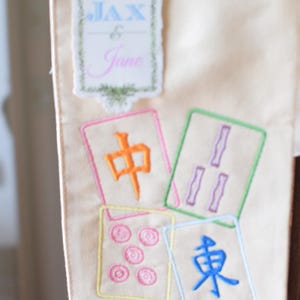 May include: Cream-colored fabric with embroidered Mahjong tiles and a decorative label that reads "Jax & Jane". The tiles feature colorful symbols and characters, perfect for game enthusiasts.