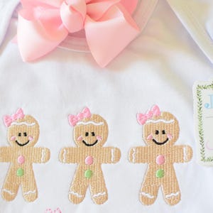May include: White onesie with three embroidered gingerbread people wearing pink bows. A pink bow is attached to the onesie. A green and white tag with the text "Jax & Jane" is attached to the onesie.