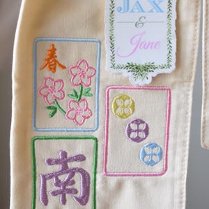 May include: Cream-colored fabric with embroidered designs. Features floral and geometric patterns in pink, yellow, purple, and blue, along with the names "Jax & Jane". The design is framed with green and blue borders.