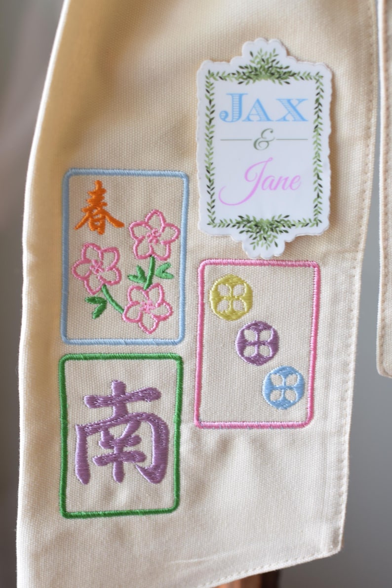 May include: Cream-colored fabric with embroidered designs. Features floral and geometric patterns in pink, yellow, purple, and blue, along with the names "Jax & Jane". The design is framed with green and blue borders.