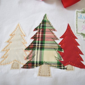 May include: White onesie with three appliqued Christmas trees. The center tree is plaid in red, green, and white. The trees are outlined in gold thread. A white tag with the text "Jax & Jane" is attached to the onesie.