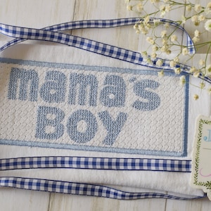 May include: White embroidered baby blanket with blue stitching that reads "mama's boy". A blue and white gingham ribbon is wrapped around the blanket. A small white tag with green foliage and the text "Jax & Jane" is in the lower right corner.