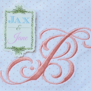 Könnte beinhalten: Ein weißer Stoff mit rosa Punkten und einem rosa gestickten Buchstaben B. Ein weißes Etikett mit grünen Blättern und dem Text "Jax & Jane" befindet sich in der oberen linken Ecke.