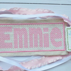 May include: A beige fabric banner with pink embroidery that reads "Emme". A small tag with green foliage and the text "Jax & Jane" is attached to the banner.