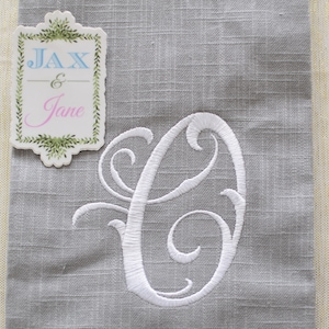 May include: A gray linen napkin with a white embroidered letter "C" and a white tag with the text "Jax & Jane" in blue and pink lettering.