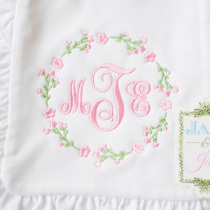 May include: White embroidered baby blanket with pink floral wreath and monogram "MJE". The blanket has a ruffled edge. A tag with the text "JAX & Jane" is attached to the blanket.