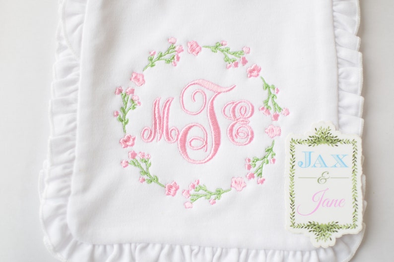 May include: White embroidered baby blanket with pink floral wreath and monogram "MJE". The blanket has a ruffled edge. A tag with the text "JAX & Jane" is attached to the blanket.