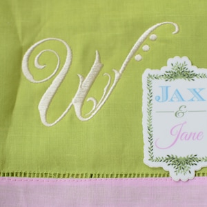 May include: A green linen tea towel with a white embroidered letter W and a white sticker with green foliage around the edges. The sticker says "Jax & Jane" in blue and pink lettering.