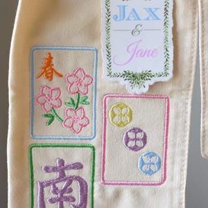 May include: Cream-colored fabric with embroidered designs. Features a sticker with the names "JAX & Jane" and floral accents. The embroidered squares include floral and Chinese character motifs in various colors.