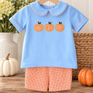 May include: A light blue short-sleeved shirt with a Peter Pan collar and orange gingham trim, featuring three embroidered orange gingham pumpkins across the chest, paired with matching orange and white gingham shorts for a coordinated set.