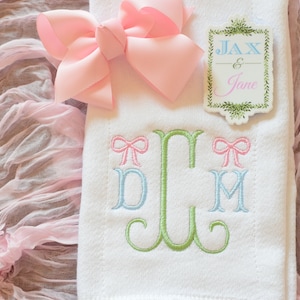 May include: A white embroidered towel with a green, blue, and pink monogram. The monogram is surrounded by two pink bows. A pink bow is on top of the towel. A white tag with green foliage and the text "Jax & Jane" is on the towel.