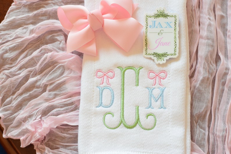 May include: A white embroidered towel with a green, blue, and pink monogram. The monogram is surrounded by two pink bows. A pink bow is on top of the towel. A white tag with green foliage and the text "Jax & Jane" is on the towel.