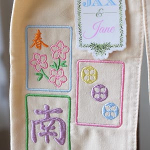 May include: Cream-colored fabric with embroidered designs. Features floral and Chinese character motifs in pink, orange, green, and purple. Includes a sticker with the names "JAX & Jane" in blue and pink.
