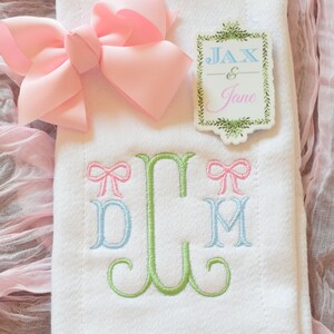 May include: White embroidered baby blanket with a green monogram of the letters "D, C, M" with pink bows on either side of the monogram. The blanket also has a pink bow and a tag that says "Jax & Jane" in green lettering.