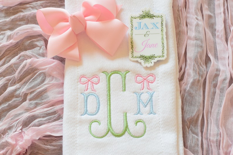 May include: White embroidered baby blanket with a green monogram of the letters "D, C, M" with pink bows on either side of the monogram. The blanket also has a pink bow and a tag that says "Jax & Jane" in green lettering.