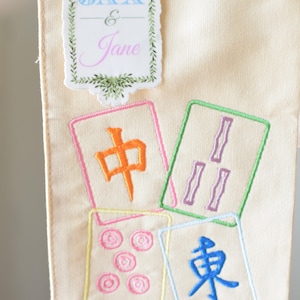 May include: Cream-colored fabric with embroidered Mahjong tiles in various colors. The tiles feature Chinese characters and symbols. A decorative label with the names "Jax & Jane" is at the top.