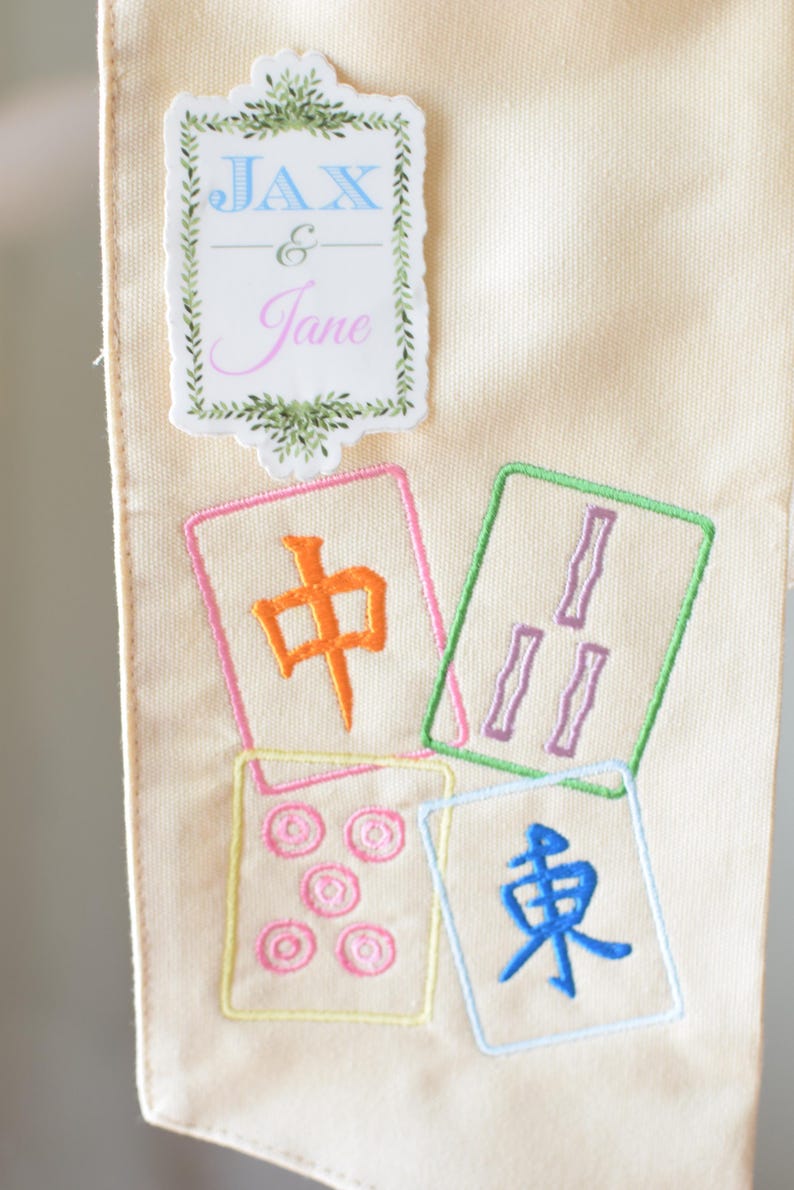 May include: Cream-colored fabric with embroidered Mahjong tiles in various colors. The tiles feature Chinese characters and symbols. A decorative label with the names "Jax & Jane" is at the top.