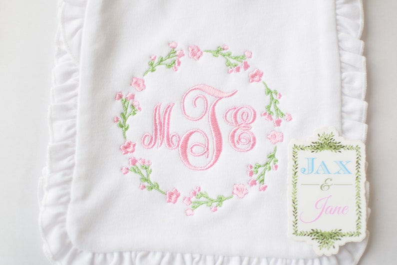 May include: White baby blanket with pink embroidered floral wreath and monogram "MJE". The blanket has a ruffled edge and a tag that reads "Jax & Jane".