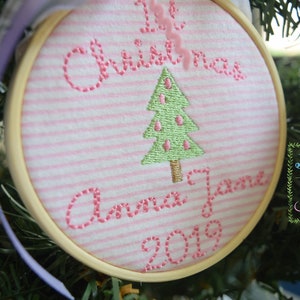 May include: A pink and white striped fabric ornament with pink embroidery that reads "1st Christmas Anna Jane 2019" and a green embroidered Christmas tree. The ornament is in a wooden hoop.
