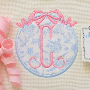 Puede incluir: Un monograma bordado con la letra rosa "J" dentro de un óvalo floral azul y blanco, coronado con un lazo rosa. También hay una etiqueta con el texto "Jax & Jane", junto con rizos de cinta rosa.