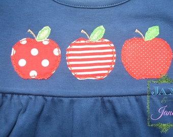Puede incluir: Una camisa azul marino con tres manzanas de fieltro rojo. Las manzanas tienen diferentes patrones: lunares, rayas y puntos pequeños. La camisa tiene un dobladillo con volantes y el texto "JAX & Jane" está bordado en la parte inferior.