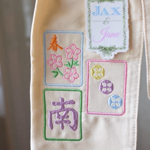 May include: Cream-colored fabric with embroidered Mahjong tile designs. The tiles feature colorful floral and geometric patterns, along with Chinese characters. A sticker with the names "Jax & Jane" is also present.