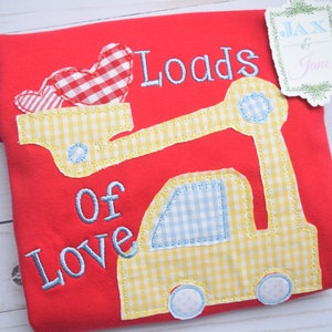 Peut inclure: T-shirt rouge avec un motif de camion de chantier jaune et bleu. Le camion transporte des cœurs en vichy rouge et blanc. Le texte "Loads of Love" est brodé sur le t-shirt.