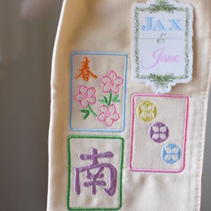May include: Cream-colored fabric with embroidered Mahjong tile designs. The designs include Chinese characters and floral patterns in pink, orange, and purple. A sticker with the names "JAX & Jane" is also visible.