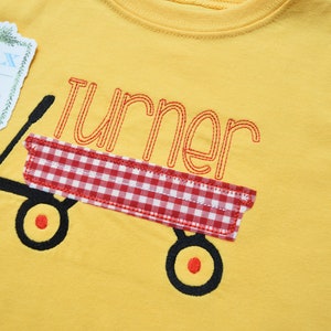 Könnte beinhalten: Gelbes T-Shirt mit einem roten und weißen karierten Wagen und dem Namen "Turner" darauf gestickt.