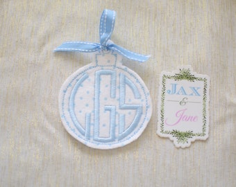 Op de afbeelding: Witte viltversiering met blauw geborduurd monogram "GS" en blauwe stippen. Een apart wit label met een groene bladrand met de tekst "Jax & Jane".