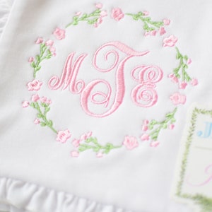 May include: White baby blanket with pink floral embroidery and a monogram in a wreath. The blanket has a ruffled edge. A tag with the text "Jax & Jane" is attached to the blanket.
