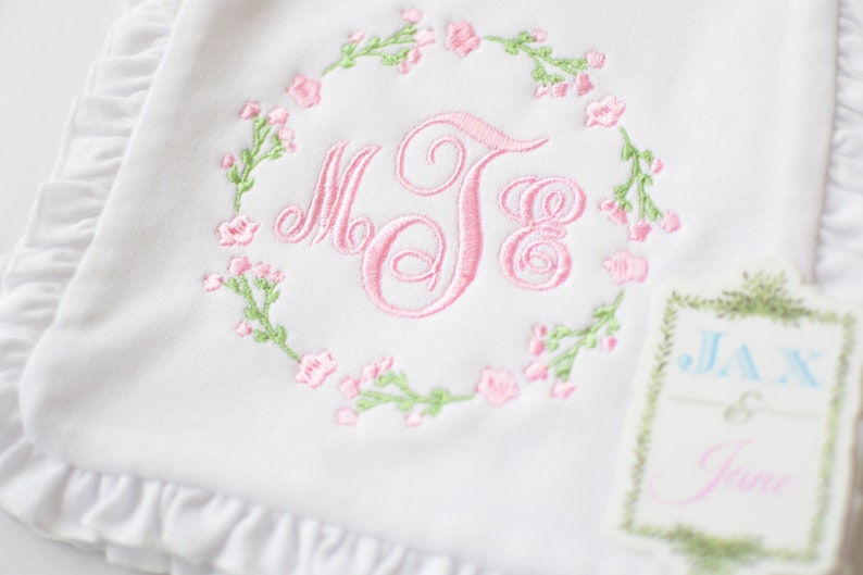 May include: White baby blanket with pink floral embroidery and a monogram in a wreath. The blanket has a ruffled edge. A tag with the text "Jax & Jane" is attached to the blanket.