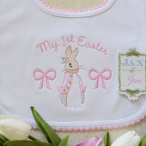May include: White bib with pink scalloped trim, embroidered with a bunny, bows, and the text "My 1st Easter". A separate tag is attached with the text "JAX & Jane" in a green leafy frame.
