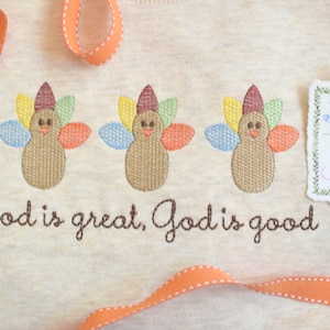 Może przedstawiać: Jasny beżowy materiał z trzema haftowanymi indykami z kolorowymi piórami. Pod indykami wyhaftowano napis "God is great, God is good". Widoczna jest również pomarańczowa wstążka i etykieta z napisem "Jax & Jane".
